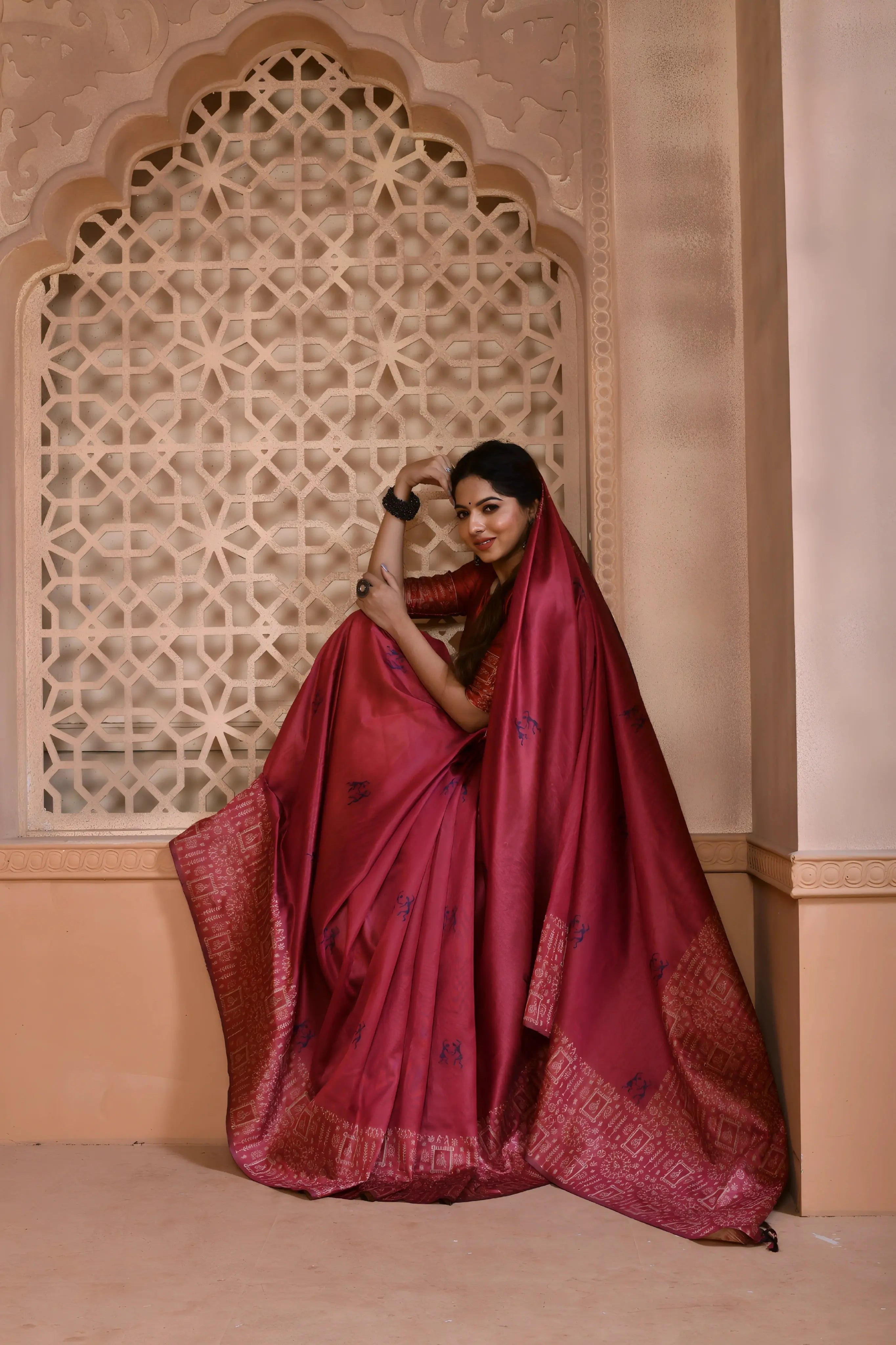 A woman models a striking Dark Pink Banglori Raw Silk Saree. The pallu and border feature dense, traditional Kalamkari-style weaving in a lighter pink/gold Zari. She wears a red blouse and substantial dark gemstone or oxidized silver jewelry, posing in an ornate, traditional setting.